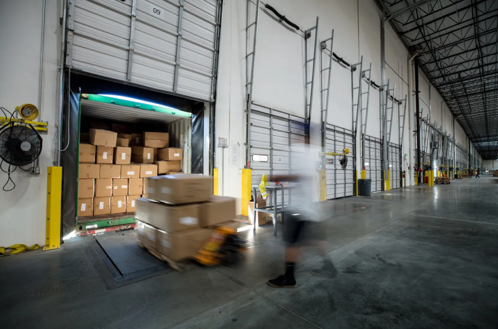 Warehouse interior with open loading dock, stacked cardboard boxes, and worker using a pallet jack, illustrating logistics and order fulfillment for ecommerce operations.
